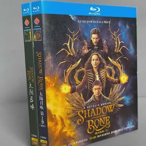 May include: Two Blu-ray disc cases for the Netflix series "Shadow and Bone" Season 2. The cases are blue and green with images of the show's characters and the series logo. The text "Every power has a price" is visible.