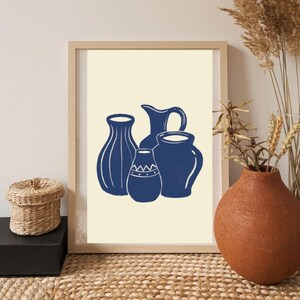 May include: A framed print featuring a collection of four blue vases in varying shapes and sizes against a cream background. The vases have white line details. The artwork is displayed on a woven surface, with a small basket and a terracotta vase in the foreground.