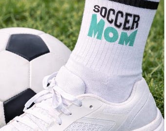Op de afbeelding: Witte sportschoen en een witte sok met zwarte strepen en de woorden "SOCCER MOM" in zwart en teal. Een voetbal staat op de achtergrond op een groen grasveld.