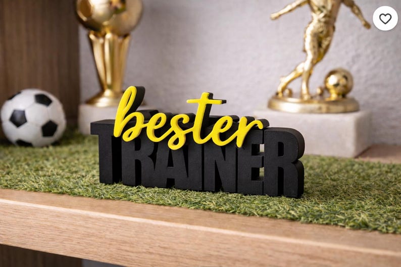 Op de afbeelding: Een zwart houten bord met de woorden "bester TRAINER" in geel en zwart. Het bord staat op een bed van groen kunstgras. Een voetbal en gouden trofee&euml;n staan op de achtergrond.