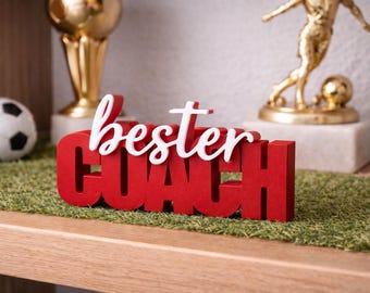 Op de afbeelding: Een rood houten bord met de woorden "bester COACH" in witte letters. Het bord staat op een groene ondergrond, met een voetbal en gouden trofeeën op de achtergrond. Een decoratief item voor een coach.