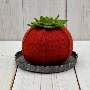 May include: A round, red felted wool tomato pincushion with a green felted leaf top, resting on a small, circular, dark gray metal tray with a scalloped edge. The tomato has visible vertical stitched seams creating a segmented, rounded shape.
