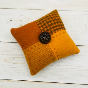 May include: A small, square, patchwork pillow with a brown button in the center. The pillow is made of four different shades of orange fabric, each with a different texture.