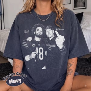 May include: A navy blue short-sleeve t-shirt featuring a vintage-style black and white graphic print of two football players. One player wears a jersey with the number 18, and the other wears a cap. The text includes signatures and the word Bears.