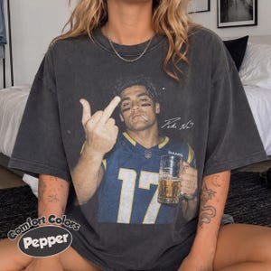 May include: A charcoal grey short-sleeve t-shirt featuring a graphic print of a football player in a blue jersey with the number 17, holding a beer mug and gesturing with a middle finger. The text "Comfort Colors Pepper" is printed in the corner.