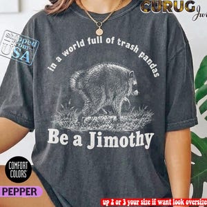 May include: A charcoal gray short-sleeve t-shirt featuring a white graphic of a raccoon. The text reads, In a world full of trash pandas Be a Jimothy. The shirt has a vintage-style washed appearance and a crew neckline.