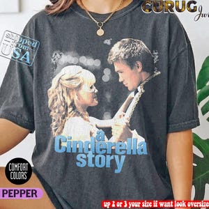 Könnte beinhalten: Ein anthrazitfarbenes Kurzarm-T-Shirt mit einem Grafikdruck einer Szene aus A Cinderella Story, auf der sich zwei Personen gegenüberstehen. Der Text A Cinderella Story ist in Hellblau unter dem Bild gedruckt. Das T-Shirt hat eine Vintage-Waschung.