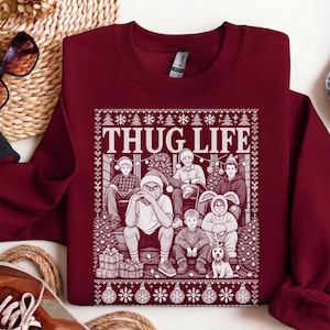 Puede incluir: Una sudadera de cuello redondo color granate con un estampado gráfico blanco de un grupo de personas y una criatura sentada en unas escaleras. El texto THUG LIFE aparece en letras blancas grandes sobre el grupo. Incluye copos de nieve.