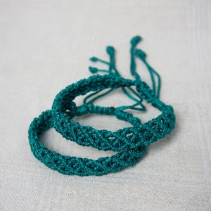 May include: Two teal macrame bracelets are displayed. The bracelets are made of woven cord, with a textured pattern. The bracelets are adjustable, with a sliding knot closure. The bracelets are on a white surface.