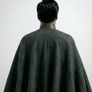 May include: A dark gray cape with a brown leather collar. The cape is made of a textured fabric and drapes over the shoulders. The person's back is facing the camera.