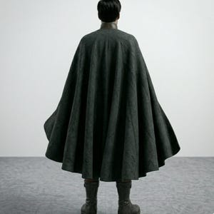 May include: A dark gray cape with a wide, flowing design. The cape is worn over a dark top and paired with matching boots. The person is seen from the back, standing on a gray tiled floor.