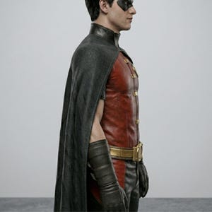 May include: A costume featuring a red and black color scheme. The outfit includes a red leather-like vest with gold accents, a black cape, and matching gloves. A gold belt cinches the waist. The character is wearing a black mask.