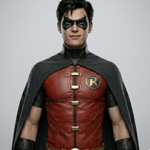 May include: A person wearing a Robin costume. The costume features a red and black leather-like bodice with gold accents and a gold "R" emblem. The person is also wearing a black cape and mask.
