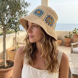 Può includere: Un cappello a secchiello all'uncinetto beige con disegni floreali blu e gialli. Il cappello ha un'ampia tesa ed è realizzato in un materiale tessuto. Un accessorio elegante per le giornate di sole.