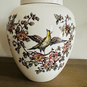 May include: Cream-colored ceramic jar with lid, adorned with a bird and floral design. The bird has yellow, brown, and black feathers, perched on a branch surrounded by pink and yellow flowers and green leaves. The jar is a decorative home accent.