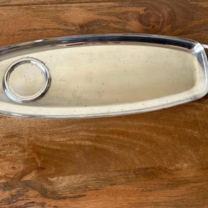 May include: An elongated, oval-shaped silver-toned metal serving tray with a polished finish. The tray features two curved handles on each end and a circular, raised-edge indentation positioned on the left side of the flat, rectangular base.