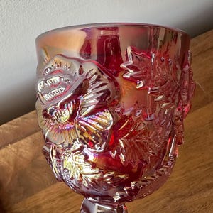 May include: A deep red carnival glass goblet featuring an iridescent finish and a raised relief pattern of blooming flowers and detailed leaves. The glass has a reflective, metallic sheen that catches the light, highlighting the intricate floral textures.