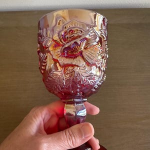 May include: A ruby-red goblet with a rose design. The glass features an embossed floral pattern, with a large rose in the center. The goblet has a round base, a faceted stem, and a wide bowl. The glass has an iridescent sheen.