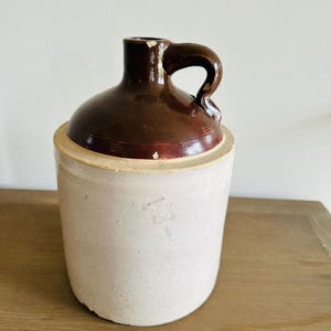 May include: A vintage stoneware jug with a brown glazed top and handle, and a cream-colored body. The jug has a rounded shape and a classic design, perfect for rustic decor.