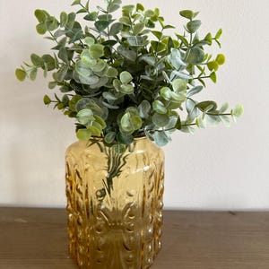 May include: A decorative arrangement featuring a golden-yellow glass vase filled with faux eucalyptus greenery. The vase has an embossed pattern. The eucalyptus has green and gray-green leaves, creating a natural, elegant display.