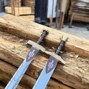 May include: Two decorative swords with bronze and gold accents rest on a wooden surface. The blades are silver with etched designs and a brown and white emblem. The hilts are dark brown with gold-colored pommels and crossguards. The text "ANAKAYΣMOΣ" is visible.