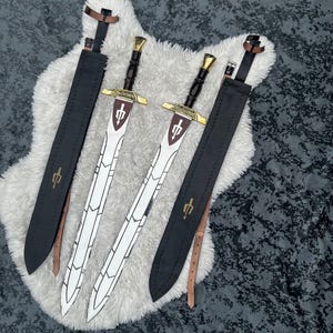 May include: Two double-edged swords with dark brown handles, gold-coloured crossguards, and silver blades featuring white geometric patterns. Each sword includes a black leather scabbard with a gold trident-like emblem and adjustable brown straps.