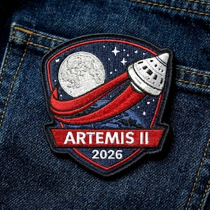 May include: Embroidered patch featuring the Artemis II mission design. The patch shows a spacecraft launching from Earth towards the moon, with the text "ARTEMIS II 2026" in red on a navy blue background.