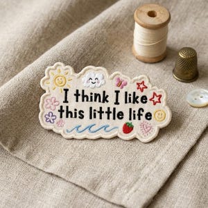 May include: An embroidered patch with the phrase "I think I like this little life" in black text, surrounded by colorful embroidered images of a sun, cloud, flowers, and a strawberry. A wooden spool of thread, a thimble, and a button are also visible.