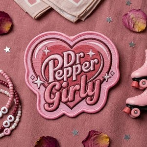 May include: A collection of pink accessories, including a heart-shaped patch with "Dr. Pepper Girly" text, a pair of roller skate keychains, beaded bracelets, and a handkerchief with cherry and heart patterns. Scattered rose petals and star-shaped confetti complete the arrangement.