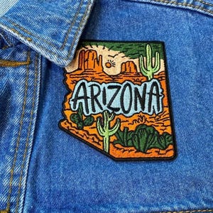 May include: Close-up of a blue denim jacket with an Arizona patch. The patch features a desert landscape with cacti, mountains, and the word "ARIZONA" in a stylized font. The jacket has a button closure and a collar.