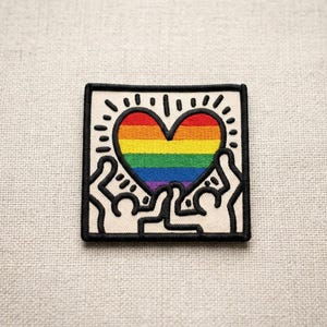 May include: Square patch with a rainbow heart design, framed in black. The heart is held by two stylized figures in black and white. The patch has a black border and is set against a neutral fabric background.