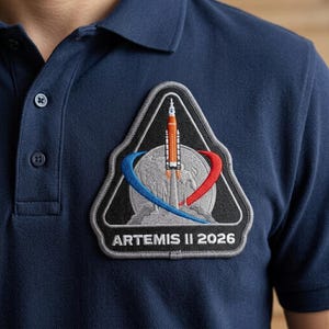 May include: Navy blue polo shirt featuring an embroidered Artemis II 2026 patch. The patch is a black triangle with a rocket launching from Earth, and blue and red swooshes. The shirt has a button-down collar.