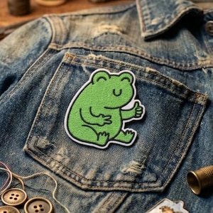 May include: A green frog patch with a white border is sewn onto the pocket of a denim jacket. The frog is giving a thumbs-up gesture. The jacket has a worn, distressed appearance with visible stitching and a button closure.