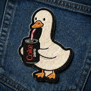 May include: Embroidered patch featuring a cartoon goose holding a Coke Zero can with a straw. The goose is white with an orange beak and feet, outlined in black. The Coke Zero can is black with red text.