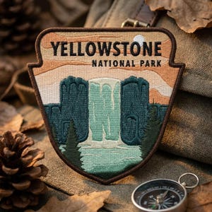 May include: A shield-shaped embroidered patch featuring the text YELLOWSTONE NATIONAL PARK in black. The design depicts a light blue waterfall cascading between dark green cliffs, with a pale orange sky and a small white moon above.