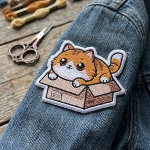 May include: Embroidered patch featuring a cartoon orange tabby cat in a cardboard box. The patch is sewn onto a denim jacket. The cat has large eyes and a white belly. Embroidery floss and scissors are in the background.