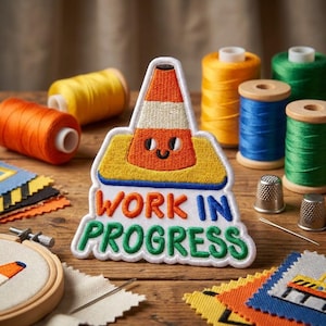 May include: An embroidered patch featuring a cartoon traffic cone with a smiling face, positioned above the text "WORK IN PROGRESS" in colorful, stitched lettering. The patch has a white border and is surrounded by sewing supplies like thread spools.