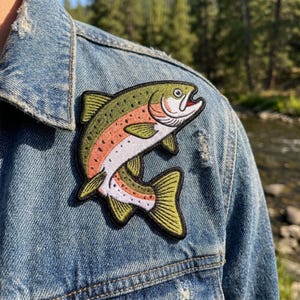 May include: An embroidered patch of a rainbow trout fish featuring olive green, pink, and white thread colors, attached to a blue denim jacket. The fish is depicted in a curved, jumping position with a black border outlining its entire shape.