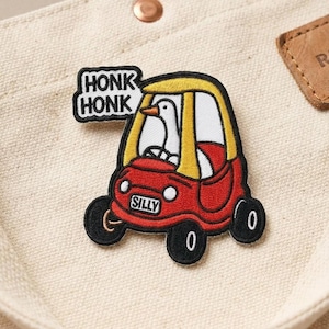 May include: Embroidered patch featuring a penguin driving a red and yellow toy car. The car has the word "SILLY" on the license plate and the text "HONK HONK" in a speech bubble. The patch is on a beige fabric.