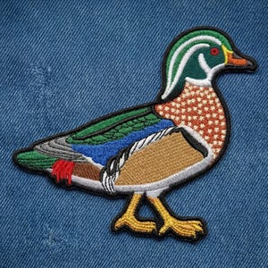 May include: Embroidered patch of a colorful duck with green, blue, brown, and orange details. The duck has a green head with white stripes, a red eye, and yellow feet. The patch is on a blue denim background.