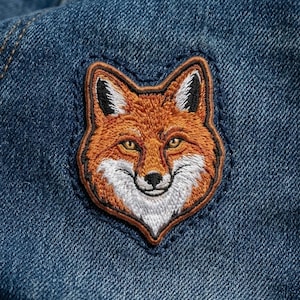May include: Embroidered patch featuring a detailed fox head with orange, white, and black accents. The patch is sewn onto a blue denim fabric, showcasing the fox's smiling face and intricate design. A decorative item for clothing or accessories.