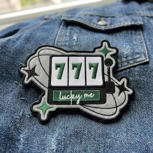 May include: Embroidered patch featuring a slot machine design with the number 777 in green. The patch has a gray and black outline with green star accents and the words "lucky me" in cursive.