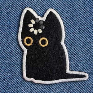 May include: Black cat patch with white embroidered outline, gold eyes, and a flower detail on its head. The patch is on a blue denim background. This embroidered patch is perfect for jackets, bags, and other accessories.