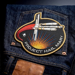 May include: A dark blue denim fabric featuring an embroidered patch with a black background. The patch displays a white spacecraft, a red curved trail, a yellow sunburst, and the white text "PROJECT HAIL MARY" along the bottom edge.