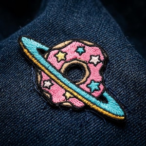 May include: Embroidered patch featuring a pink planet with a donut-like shape, adorned with star-shaped accents in yellow, white, and blue. The planet has a light blue ring with a yellow stripe, set against a dark blue denim background.