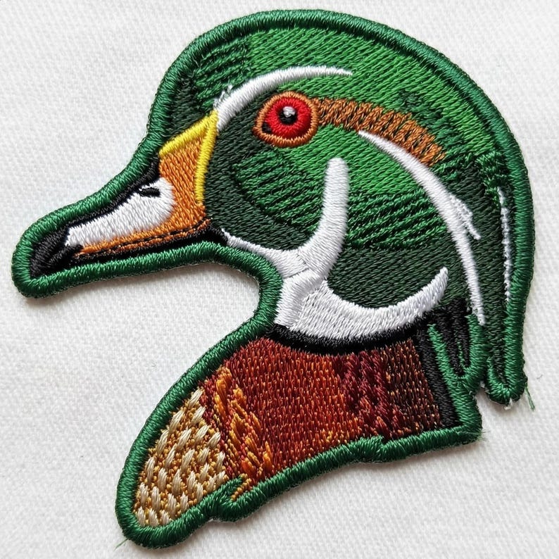 May include: Embroidered patch featuring a detailed depiction of a duck's head. The patch showcases a vibrant color palette, including shades of green, brown, orange, yellow, white, and red. The duck's eye is a striking red, and the design is outlined with a green border.