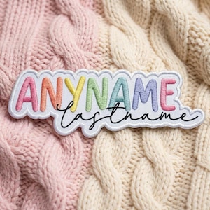 May include: A white embroidered patch with a scalloped edge featuring the word ANYNAME in colorful, puffy, pastel-toned letters and the word lastname written below in a black, cursive, handwritten-style script font.