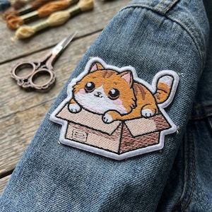 May include: Embroidered patch featuring a cartoon orange tabby cat in a cardboard box. The patch is sewn onto a denim jacket. The cat has large eyes and a white belly. Embroidery floss and scissors are in the background.
