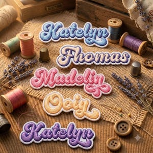 May include: Five embroidered name patches with a retro script font, featuring the names Katelyn in blue, Thomas in purple, Madelyn in pink, Oily in yellow, and Katelyn in purple, arranged on a textured fabric surface with sewing supplies.