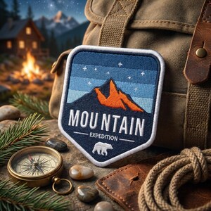 May include: A shield-shaped embroidered patch featuring a dark blue mountain range with an orange peak under a starry sky. The text reads MOUNTAIN EXPEDITION in white capital letters, accompanied by a small white bear silhouette at the bottom.